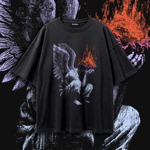 Puede incluir: Una camiseta gris carbón de manga corta con un estampado gráfico de una figura alada agachada con la cabeza entre las manos. La figura está representada en tonos morados y blancos, con un efecto de llamas naranja y rojo sobre su cabeza.