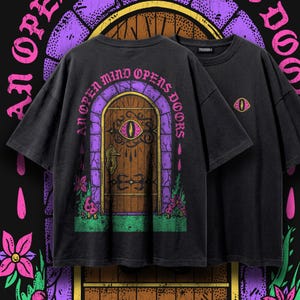 May include: A black t-shirt featuring a graphic print of a wooden door with an eye in the center, surrounded by a purple stone archway, flowers, and mushrooms. The text "AN OPEN MIND OPENS DOORS" is printed in pink. A small eye icon is on the chest.