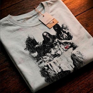 Puede incluir: Camiseta gris claro con un estampado gráfico en blanco y negro de varias figuras. El diseño tiene un estilo artístico y desgastado. Una etiqueta marrón está sujeta a la camiseta.