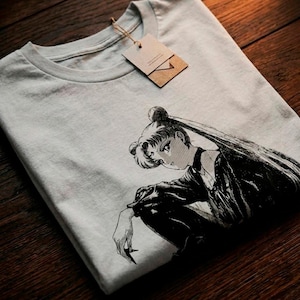 Puede incluir: Una camiseta gris claro con una ilustración en blanco y negro de estilo anime de una mujer con pelo largo. La camiseta tiene cuello redondo y una etiqueta adjunta con una cuerda. La camiseta está doblada sobre una superficie de madera.