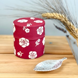 May include: A red cylindrical box with a white floral pattern and a silver leaf-shaped tea scoop. The box is on a wooden surface.