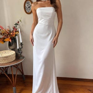 May include: A floor-length, strapless white gown featuring a fitted bodice and a smooth, satin-like finish. The dress has a clean, minimalist silhouette that flows straight down to the floor, creating a simple and elegant appearance.