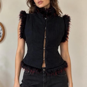 May include: A sleeveless black vest featuring a high collar and trim made of black faux fur with subtle red highlights. The garment closes at the front with four silver-toned metal hook-and-eye fasteners, leaving a small vertical gap between the panels.