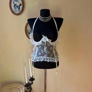 May include: A white lace halter top featuring a sheer floral pattern, ruffled trim along the neckline and hem, and a small center ribbon bow. The garment includes two thin dangling lace straps at the bottom and is displayed on a black mannequin.