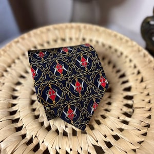 May include: Navy blue tie with a repeating pattern of red and white shapes, outlined in gold, on a dark background. The tie is displayed on a woven, light brown surface.