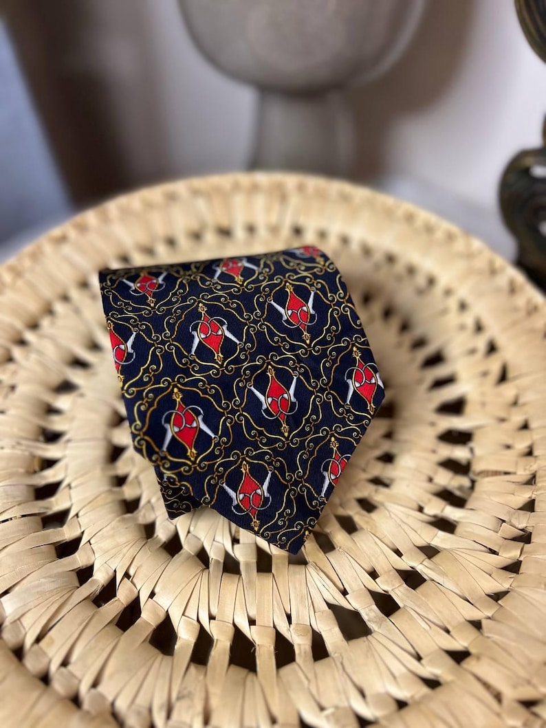 May include: Navy blue tie with a repeating pattern of red and white shapes, outlined in gold, on a dark background. The tie is displayed on a woven, light brown surface.