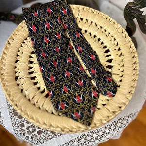 May include: A navy blue tie with a repeating pattern of red and white heraldic symbols and gold accents. The tie is displayed on a woven straw basket. The tie's design is a classic and elegant style.
