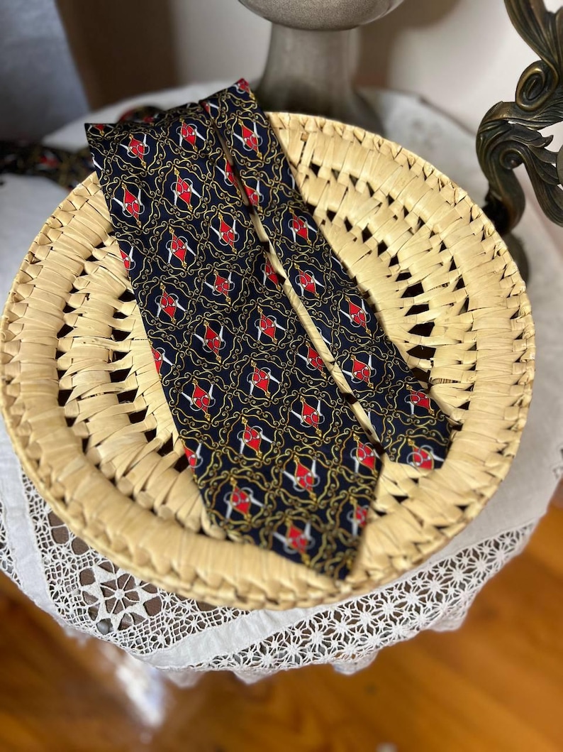May include: A navy blue tie with a repeating pattern of red and white heraldic symbols and gold accents. The tie is displayed on a woven straw basket. The tie's design is a classic and elegant style.