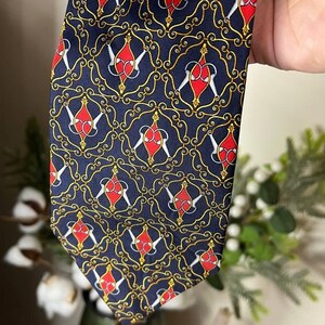 May include: Navy blue tie with a repeating pattern of red and white shapes within gold scrollwork. The tie is held up, showcasing the intricate design and color contrast. A classic accessory.