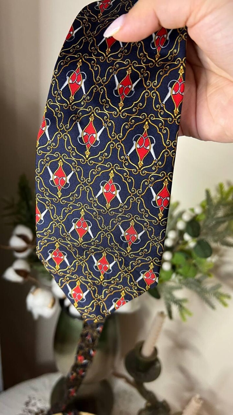 May include: Navy blue tie with a repeating pattern of red and white shapes within gold scrollwork. The tie is held up, showcasing the intricate design and color contrast. A classic accessory.