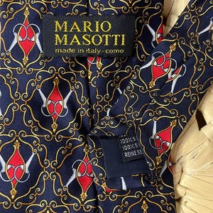 May include: Navy blue silk tie with a repeating pattern of red and white shapes, outlined in gold, on a navy background. The label reads "MARIO MASOTTI made in italy - como".