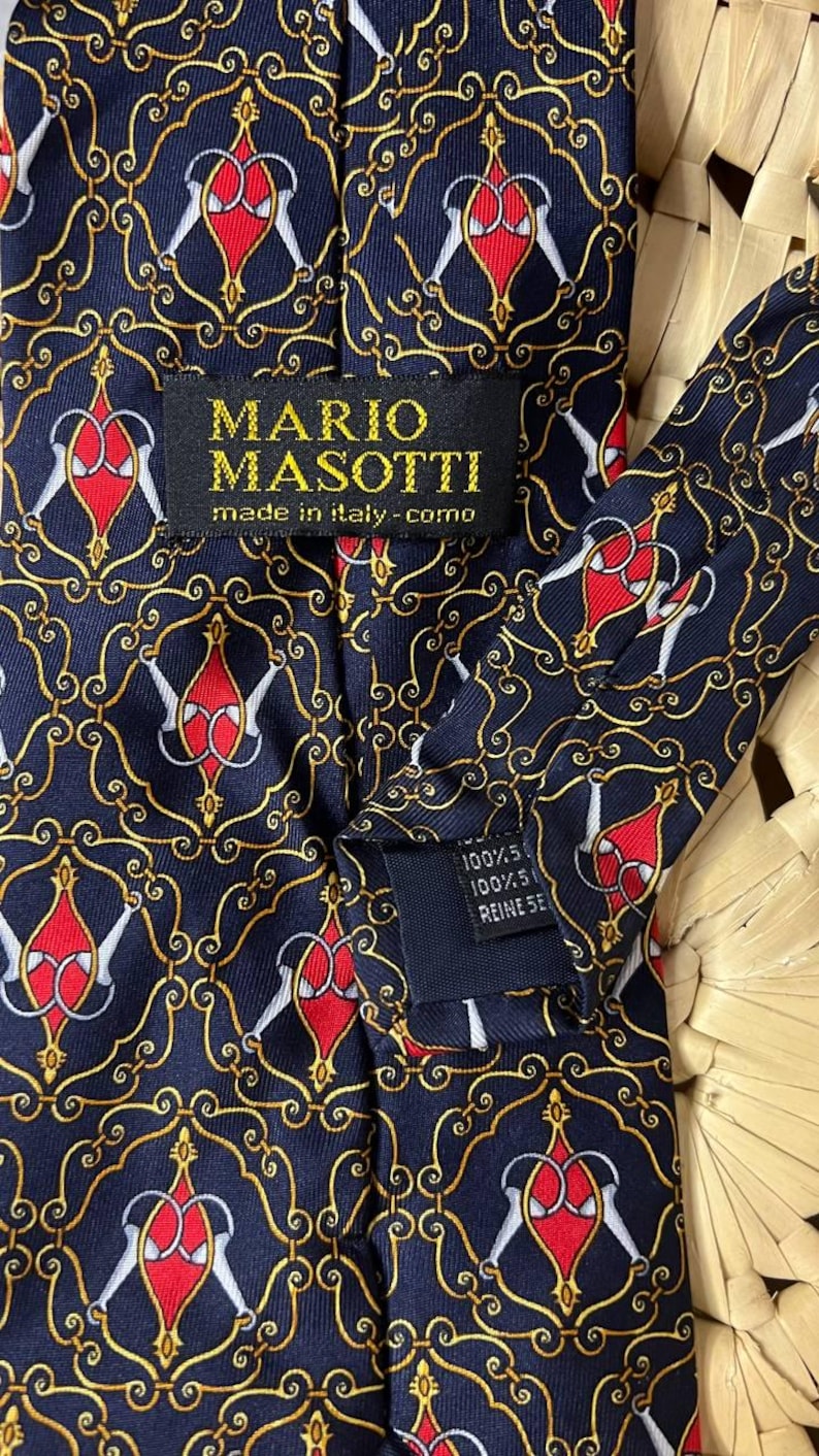 May include: Navy blue silk tie with a repeating pattern of red and white shapes, outlined in gold, on a navy background. The label reads "MARIO MASOTTI made in italy - como".
