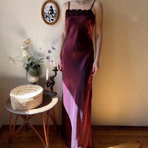 May include: A floor-length, deep burgundy slip dress featuring thin spaghetti straps and a row of matching fabric rosettes along the straight neckline. The garment is crafted from a shimmering, satin-like fabric that reflects light.