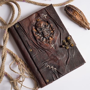 Op de afbeelding: Een bruin leren dagboek met een gegraveerde schedel op de voorzijde. Het dagboek heeft een metalen sluiting en is versierd met stiksels. Het dagboek ligt op een wit oppervlak met een stuk touw en een gedroogde zaadpeul.