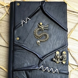 Op de afbeelding: Zwart lederen dagboek met een drakenontwerp en een metalen sluiting. Het dagboek heeft een verweerde look en is gestikt met wit garen.