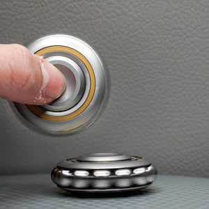 May include: A silver fidget spinner with gold detailing is held by a finger, with another spinner below. The top spinner is in motion, highlighting its design. The background is a textured grey surface.
