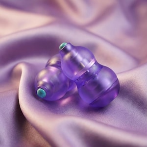 May include: Two translucent purple earplugs with teal tips rest on a lavender satin surface. The earplugs have a gourd-like shape. The background is a soft, blurred gradient of lavender and purple.