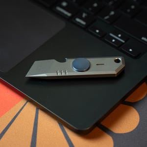 Puede incluir: Un pequeño cuchillo utilitario plateado con un botón giratorio azul oscuro y un abridor de botellas. El cuchillo está apoyado sobre un panel táctil negro de un portátil, con un teclado al fondo. El fondo incluye una superficie naranja y negra estampada.