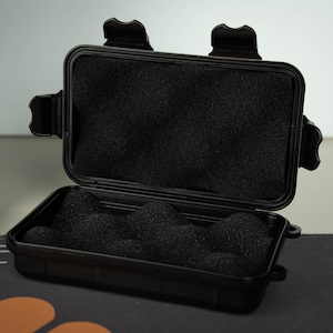 Puede incluir: Una pequeña caja negra abierta con interior de espuma. La caja tiene forma rectangular con esquinas redondeadas y una tapa con bisagras. La espuma interior es negra y tiene una superficie texturizada. La caja está sobre una superficie estampada.