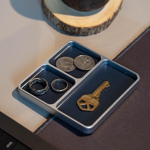 May include: A silver and navy blue tray with four compartments. The tray holds two rings, two coins, and a gold key. The tray is on a desk with a keyboard and a wooden coaster in the background. The tray is designed to hold small items.