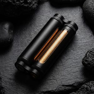 May include: A black, cylindrical device with gold accents and a textured pattern, resting on a dark, textured surface. The device has a sleek, modern design, with a gold-colored, grid-like pattern visible through a clear window. The background features dark, rough-textured stones.