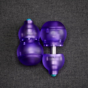 Puede incluir: Un juego de dos tapones de botella de vidrio morado con un conector de metal plateado. Los tapones tienen una forma redondeada y bulbosa con un pequeño acento turquesa en la parte superior.