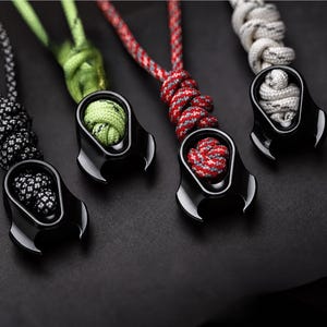 May include: Four black metal zipper pulls, each featuring a unique paracord knot. The cords are black with white speckles, lime green, red with grey patterns, and white with grey speckles. Each pull has a smooth, curved, claw-like design.