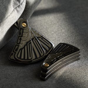 Puede incluir: Un juego de tres navajas plegables de bolsillo de metal negro con intrincados patrones geométricos. Las navajas tienen detalles dorados y están plegadas.