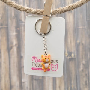 May include: Orange and pink cat keychain with a silver chain and a key ring. The keychain is hanging from a wooden clothespin on a rope. The background is a wooden surface. The text on the card reads "Meow-gical Treasures Jewelry & Accessories".