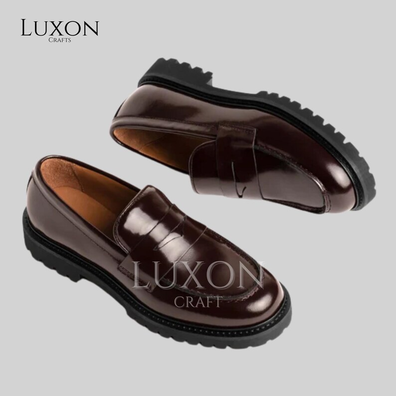 Może przedstawiać: Dwa brązowe mokasyny penny z czarnymi podeszwami. Buty mają błyszczące wykończenie i klasyczny design. Marka "LUXON CRAFTS" jest widoczna na butach.