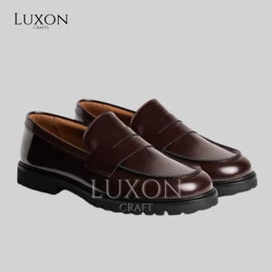 Może przedstawiać: Para ciemnobrązowych sk&oacute;rzanych mokasyn&oacute;w z błyszczącym wykończeniem. Buty mają klasyczny design penny loafer z zaokrąglonym noskiem i grubą czarną podeszwą. Widoczna jest nazwa marki "LUXON CRAFTS".