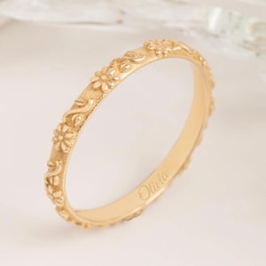 以下が含まれることがあります： 金色のリング。リングには、小さな花と葉がバンドの周りに繰り返されるパターンがあります。「Olivia」という名前がリングの内側に刻まれています。リングは柔らかい白い背景に配置されています。