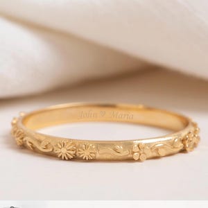 Könnte beinhalten: Ein goldfarbener Ring mit floralen und Blattdetails. Der Ring ist mit den Namen "John & Maria" graviert. Das Bild zeigt Diagramme des Ringdesigns und der Geburtsblumenoptionen, darunter Weißdorn und Gänseblümchen.