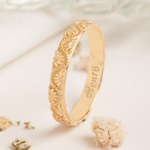 May include: A gold ring featuring a floral design and the name "Henry" engraved inside. The ring's surface is adorned with a repeating pattern of small flowers. The background is a soft, neutral colour.
