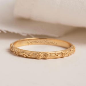 Może przedstawiać: Złoty pierścionek z grawerowanym wzorem kwiatowym i napisem "Demie & Thomas". Pierścionek ma ciągły wzór małych kwiatów i płynących linii wokół obrączki.