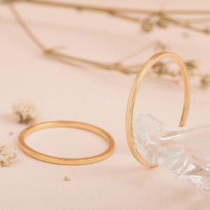 Puede incluir: Dos anillos dorados sobre una superficie rosa claro. Un anillo está plano, el otro está apoyado contra un material translúcido. Flores secas están esparcidas alrededor de los anillos.