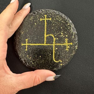 Peut inclure: Un dessous de verre circulaire en ardoise noire orné d'un symbole alchimique de Saturne peint en or au centre. La surface du dessous de verre est parsemée de taches de peinture dorée, créant un aspect texturé sur la base en pierre mate.
