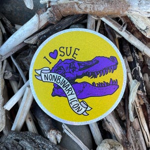 May include: A round sticker with a yellow background and a purple dinosaur skull. The text "I ♡ SUE" is above the skull, and a banner below reads "NONBINARY ICON". The sticker has a silver glitter border.