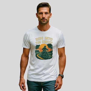 Könnte beinhalten: Weißes T-Shirt mit einer Grafik eines Motorradfahrers auf einer kurvenreichen Straße, mit Sonnenuntergang und Meereswelle im Hintergrund. Der Text "RIDE INTO THE SUNSET" ist über dem Bild gedruckt.