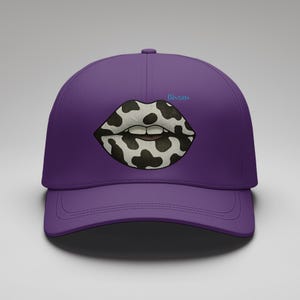 Op de afbeelding: Een paarse baseballcap met een afbeelding van lippen in een zwart-wit koeienprint. Het woord "Bisous" staat in lichtblauw boven de lippen. De cap heeft een gebogen klep en een gestructureerde kroon.