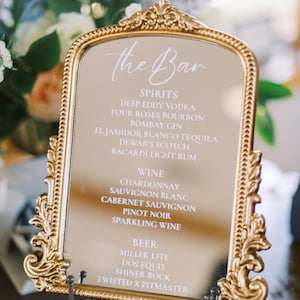 Puede incluir: Espejo con marco dorado con una lista de bebidas para un bar. El texto "the Bar" está en la parte superior, seguido de categorías de licores, vinos y cervezas. El espejo está sostenido por un soporte de metal decorativo.