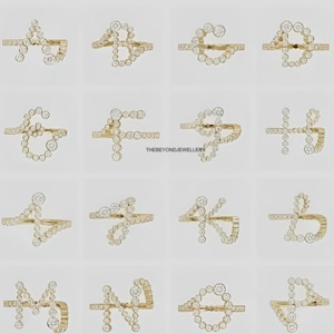 Puede incluir: Una colección de anillos dorados, cada uno diseñado en forma de letra del alfabeto. Los anillos están adornados con pequeñas piedras preciosas transparentes. El fondo es de un color claro y neutro. El texto "THEBEYONDJEWELLERY" es visible.