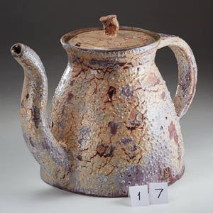May include: A ceramic teapot with a brown, white, and purple crackle glaze. The teapot has a lid and a spout. The teapot is sitting on a white surface with the numbers 1 and 7 visible in the foreground.