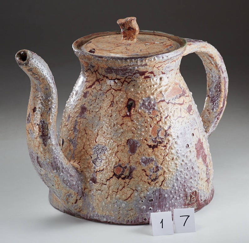 May include: A ceramic teapot with a brown, white, and purple crackle glaze. The teapot has a lid and a spout. The teapot is sitting on a white surface with the numbers 1 and 7 visible in the foreground.