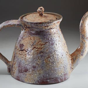 May include: A ceramic teapot with a light brown, white, and purple glaze. The teapot has a rounded body and a spout that curves up and out. The handle is attached to the side of the teapot and curves up and over the top.