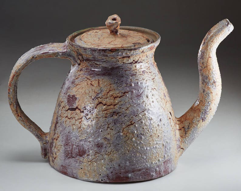 May include: A ceramic teapot with a light brown, white, and purple glaze. The teapot has a rounded body and a spout that curves up and out. The handle is attached to the side of the teapot and curves up and over the top.