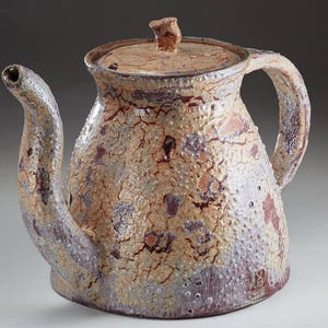 May include: A ceramic teapot with a light brown, white, and purple glaze. The teapot has a rounded body with a wide spout and a handle. The teapot has a lid with a small knob on top. The teapot has a crackled glaze and a small mark on the bottom.