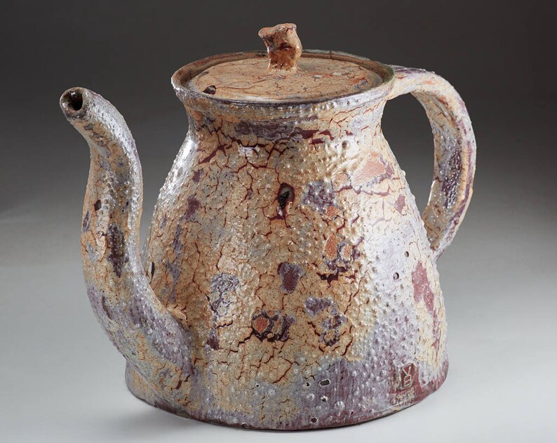 May include: A ceramic teapot with a light brown, white, and purple glaze. The teapot has a rounded body with a wide spout and a handle. The teapot has a lid with a small knob on top. The teapot has a crackled glaze and a small mark on the bottom.