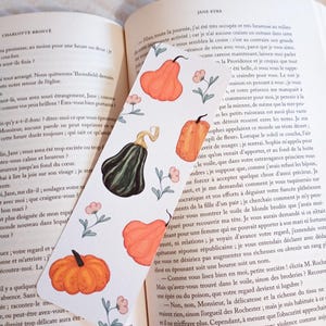 Peut inclure: Un marque-page rectangulaire en papier à fond blanc orné d'un motif répétitif de citrouilles orange, d'une courge vert foncé et de petites fleurs roses aux tiges vertes, posé sur les pages ouvertes du livre Jane Eyre.