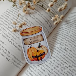 Peut inclure: Un autocollant représentant un bocal avec l'inscription "AUTUMN MEMORIES" et des illustrations de citrouilles. Les citrouilles sont orange et blanches avec des nœuds noirs. L'autocollant est sur un livre ouvert avec des fleurs séchées.